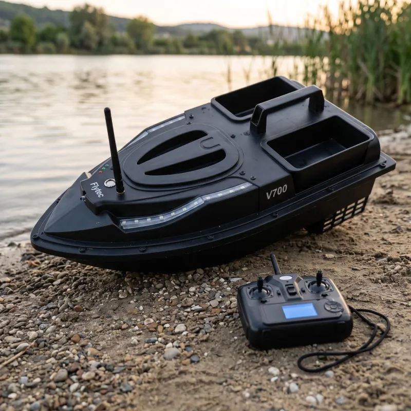 Матово черна лодка за захранка Flytec V700 с двойни бункери на брега на езеро, с антена и дистанционно управление наблизо